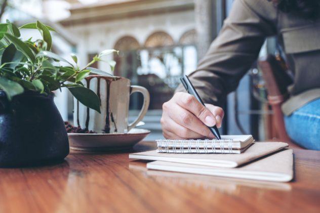 Closeup image of a woman's hand writing on blank notebook with coffee cup on wooden table Cultivating determination and consistency in book writing - How to stay motivated throughout the book writing journey - Turning writing aspirations into tangible accomplishments - The role of determination and consistency in successful authors 